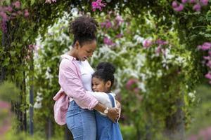 afrikansk mor och dotter kramas varje Övrig medan lyckligt gående under de blomma galler båge i de offentlig parkera under sommar för familj kärlek och vård i mors dag firande foto