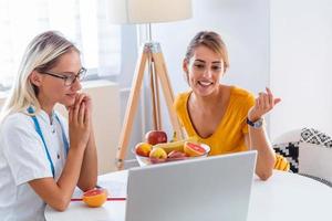 läkare nutritionist, dietist och kvinna patient på samråd i de kontor. ung leende kvinna dietist i de samråd rum. dietist skrivbord med friska frukt och mätning tejp. foto