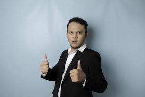 upphetsad asiatisk affärsman bär kostym ger tummen upp hand gest av godkännande, isolerat förbi blå bakgrund foto