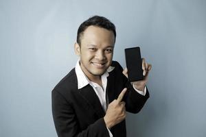 en porträtt av en Lycklig asiatisk affärsman är leende och innehav hans smartphone som visar kopia Plats på dess skärm bär svart kostym isolerat förbi en blå bakgrund foto