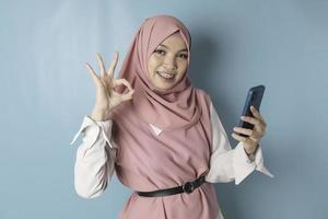en porträtt av en Lycklig asiatisk muslim kvinna är leende och innehav henne smartphone och gestikulerar ok tecken foto