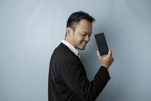 en porträtt av en Lycklig asiatisk affärsman är leende och innehav hans smartphone som visar kopia Plats på dess skärm bär svart kostym isolerat förbi en blå bakgrund foto