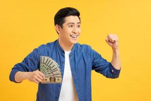 framgångsrik asiatisk man. Lycklig ung man innehav pengar medan stående och vapen upp, isolerat på gul bakgrund foto