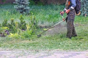 en verktyg arbetstagare klipper en grön gräsmatta med en bensin trimmer nära en fotgängare trottoar på en sommar dag. foto
