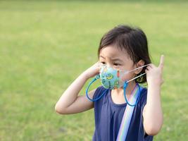 dramatisk porträtt av 4 år gammal söt bebis asiatisk flicka, liten preschooler barn med förtjusande kort hår med orolig uttryck tar av skyddande ansikte mask stående utomhus- foto