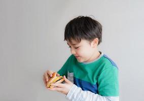 friska ung pojke äter hamburgare. en barn innehav ostburgare och ser med leende ansikte, isolerat Lycklig unge har snabb mat hamburgare för mellanmål. favorit barns mat. foto