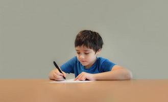 barn som sitter på bordet och gör läxor, barnpojke håller svart penna och skriver på vitt papper, ung pojke som tränar engelska ord hemma. grundskola och hemundervisning, distansundervisning koncept foto