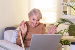 Lycklig mogna kvinna vinka till någon medan har en video ring upp över bärbar dator på Hem. gråhårig senior kvinna vinka hand i främre av bärbar dator medan har video ring upp med henne familj medlemmar. foto