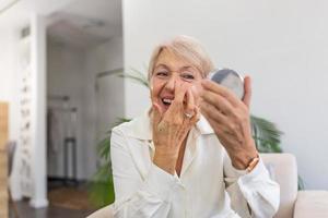 leende senior kvinna applicering anti-åldring lotion till ta bort mörk cirklar under ögon. Lycklig mogna kvinna använder sig av kosmetisk grädde till Dölj rynkor Nedan ögon. lady använder sig av dag fuktighetskräm. foto