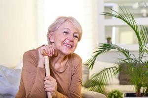 äldre lady Sammanträde på de soffa med trä- gående pinne och leende. Lycklig äldre kvinna avkopplande på soffa och innehav gående pinne. kopia Plats. senior kvinna ser omtänksam i en pensionering Hem foto