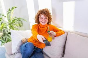 ung kvinna med lockigt röd hår innehav avlägsen kontrollera och tittar på tv, ändring kanaler. söt flicka dricka orange juice och tittar på TV i henne lägenhet foto