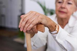 händer av ett gammal kvinna med en sockerrör, äldre lady Sammanträde på de soffa med trä- gående pinne. beskurna skott av en senior kvinna innehav en sockerrör i en pensionering Hem foto