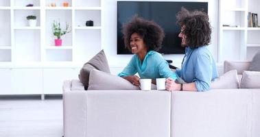 multietnisk par Sammanträde på soffa på Hem dricka coffe foto