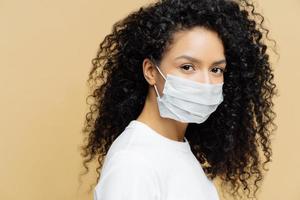 Foto av mörk flådd afro amerikan kvinna med lockigt buskig hår, bär skyddande mask under coronavirus utbrott, isolerat på beige bakgrund. förhindra sjukdom. virus, influensa, hälsa vård