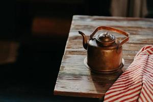 gammal kopparmetalltekanna på träbord i mörkt rum. rödrandig handduk i närheten. antik vattenkokare för att göra te eller kaffe. matlagningsutrustning foto