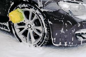 beskuren bild av svart bil tvättad av servicearbetare vid biltvätt. fordon täckt med skumbubblor. fokus på hjulet. foto