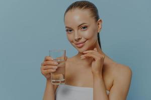frisk och återfuktad hud. ung charmig kvinna som håller ett glas rent vatten och rör vid ansiktshuden foto