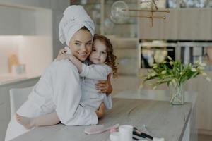 foto av ung mor och dotter omfamnar och uttrycker kärlek till varandra poserar i modern lägenhet bär mjuka vita badrockar som kommer att ha kosmetiska ingrepp har frisk hud. tid för skönhet