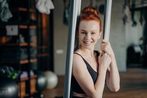 upphetsad rödhårig kvinna som njuter av antennyoga i fitnessstudio foto