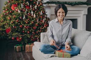 upprymd italiensk kvinna med slutna ögon som försöker packa upp julklappen när hon sitter på soffan hemma foto