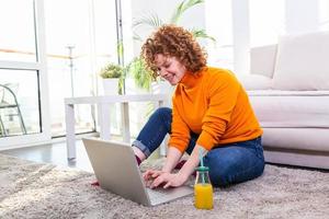 bild av en ung röd hår kvinna dricka orange juice och surfing i de internet på Hem. frilansare kvinna arbetssätt på bärbar dator från Hem foto