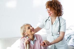 Lycklig patient är innehav vårdgivare för en hand medan utgifterna tid tillsammans. äldre kvinna i amning Hem och sjuksköterska. åldrig elegant kvinna på amning Hem foto