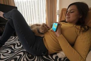 skön ung latinamerikan kvinna liggande på henne säng som visar en katt video på henne smartphone till henne liten ljus brun kattunge foto