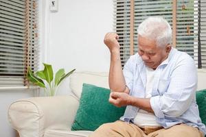 ett äldre asiatisk man sitter med ärm smärta på de soffa i hans hus. begrepp av behandling av artros, åldring sjukdom. kopia Plats foto