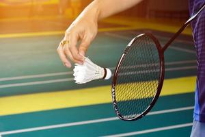 badminton racket och gammal vit fjäderboll innehav i händer av spelare medan tjänande den över de netto ett huvud, fläck badminton domstol bakgrund och selektiv fokus foto