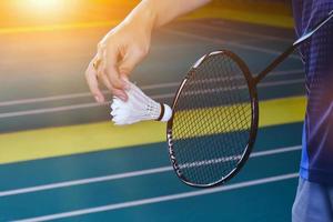 badminton racket och gammal vit fjäderboll innehav i händer av spelare medan tjänande den över de netto ett huvud, fläck badminton domstol bakgrund och selektiv fokus foto