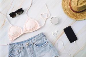 platt lägga av sommar objekt med pastell bikini, solglasögon, jeans, hatt, smink, hatt och smartphone på marmor bakgrund, mode och sommar begrepp foto