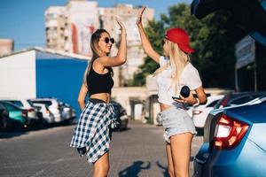 två ung flickor i solglasögon Framställ för de kamera på de bil parkering. foto
