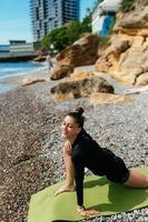 ung smal kvinna håller på med övning på yoga matta utomhus på sten strand förbi de hav foto