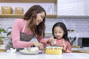 Lycklig asiatisk mor undervisning henne ung dotter till dekorera deras hemlagad kaka i vit modern kök för Hem laga mat och bakning begrepp foto