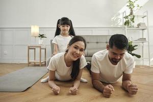 lycklig välbefinnande asiatisk thai familj, barn leker och retar sina föräldrar medan yoga fitnessträning och hälsoträning tillsammans i det vita vardagsrummet, inhemsk livsstil, helgaktivitet. foto