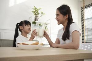 en friska asiatisk thai familj, liten dotter, och ung mor dryck färsk vit mjölk i glas och bröd glädje tillsammans på en dining tabell i morgon, wellness näring Hem frukost måltid livsstil. foto