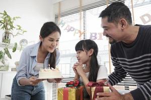 Lycklig asiatisk thai familj, ung dotter är överraskad, födelsedag kaka och härlig gåva, flicka slag ljus, be och glad firar fest med föräldrar tillsammans, särskild inhemsk Hem händelse livsstil. foto