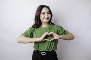 ett attraktiv ung asiatisk kvinna bär en grön t-shirt känner Lycklig och en romantisk former hjärta gest uttrycker anbud känslor foto