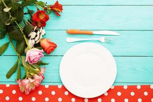 festlig middag med bestickkniv, gaffel, tallrik och rosor på blå träbakgrund. toppvy. håna. blommor. kopieringsutrymme foto