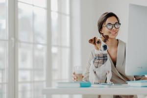 foto av kvinna som jobbar frilans hemifrån, koncentrerad i datorskärm, bär glasögon, poserar vid skrivbordet med jack russel terrier hund, dricker dryck, ler positivt tycker om favoritarbete