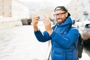 snygg man med blå charmiga ögon och skägg klädd i blå anorak, glasögon och keps gör selfie med mobiltelefon mot storstadsbakgrund. ung turist med ryggsäck gör foto av sig själv