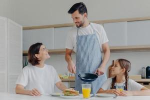 glad far, mor och dotter samlas i köket, äter utsökt frukost, pappa lagade stekt ägg, är på gott humör, redo att börja en ny dag. härlig familj njut av välsmakande måltid hemma foto