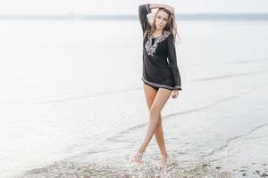 Söt smal friska ung kvinna med lång smal ben står nära lugna hav med liten vågor och blå bakgrund, varelse nöjd till utgör på kamera, känner självsäker, beundrar skön marinmålning foto