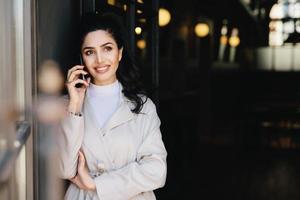 elegant brunett affärskvinna med mörka ögon och fylliga läppar klädd i vita kläder som kommunicerar över telefon med ett charmigt leende som tittar upp med ett eftertänksamt uttryck. teknik koncept foto