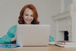 fokuserad glad rödhårig framgångsrik affärskvinna forskar, gör affärsprojekt, sitter framför bärbar dator på hemmakontorets skrivbord, använder programvara, har ett glatt leende på läpparna foto