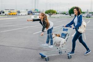 foto av glad liten flicka med sin mamma och husdjur står i kundvagnen, shoppar under helgen, njuter av fritiden, återvänder hem med nya inköp. familj, fritid, djur och inköpskoncept