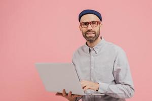 foto av kille som internetanvändare surfar på webbplatsen på bärbar dator, ansluten till trådlöst internet, håller elektronisk pryl, bär glasögon, formell skjorta, isolerad över rosa bakgrund med ledigt utrymme
