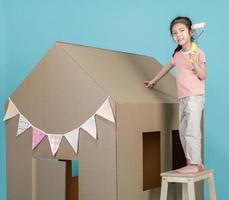 asiatisk liten barn flicka målning henne kartong hus isolerat på blå bakgrund, kreativ på Hem med familj begrepp foto