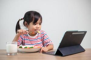 förtjusande Asien barn flicka har lunch medan tittar på en film från de läsplatta. en liten asiatisk barn äter middag och ögon är ser tecknad serie från läsplatta. nationell äter störningar medvetenhet vecka. foto