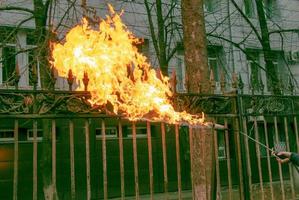 en arbetstagare rengör gammal måla från en metall staket använder sig av brand. en mannens hand innehar en gas brännare med en belyst flamma foto